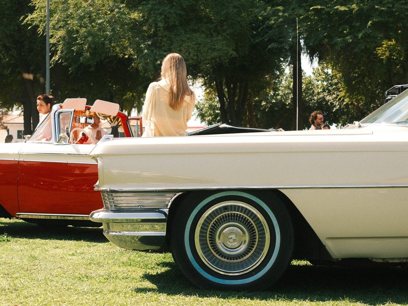 A classic white car with a prominent wheel in the foreground, vintage cars and people in the background.