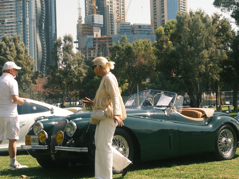 A classic green convertible car parked on grass with a woman in white pants and a light top nearby in an urban park.