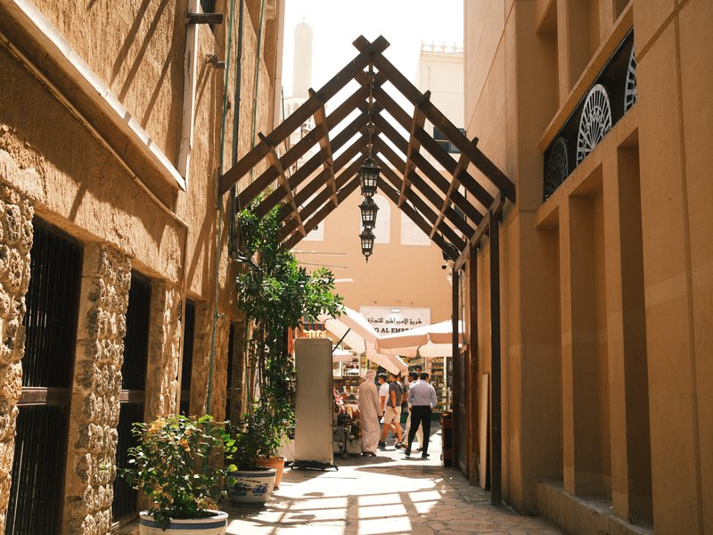 A narrow alleyway with wooden beams and plants, leading to outdoor seating areas in a sunny setting.