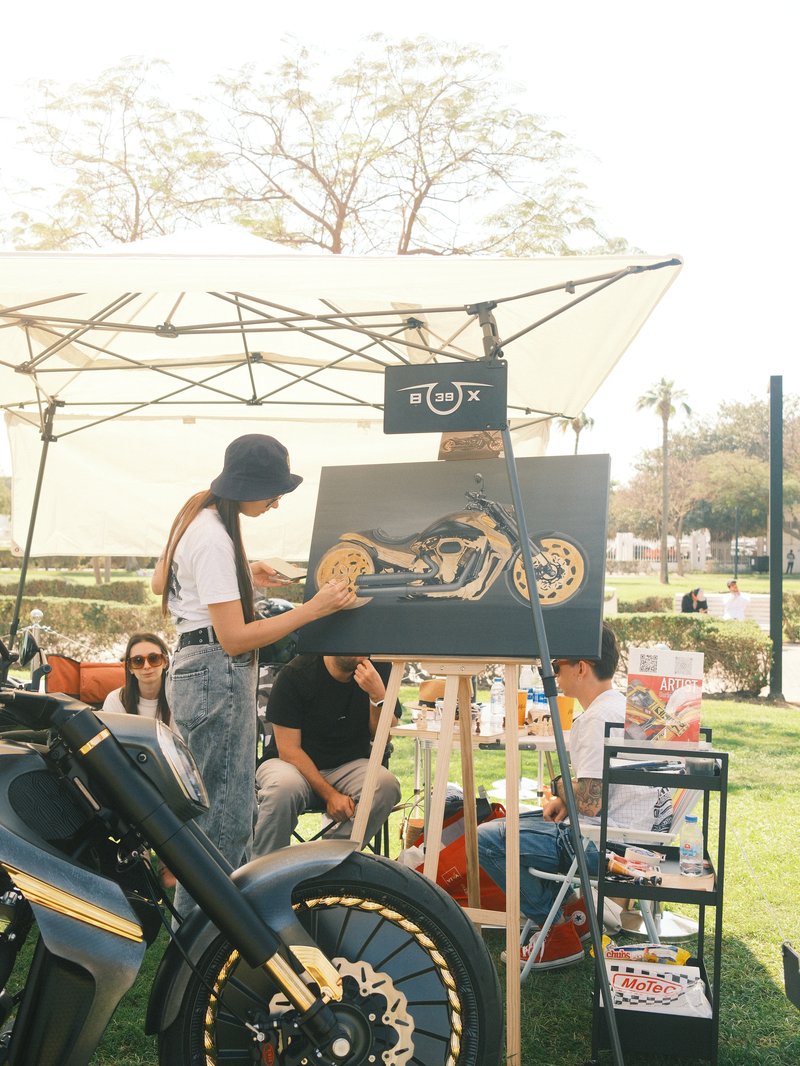 A woman painting a motorcycle on canvas at an outdoor art event with spectators in the background.