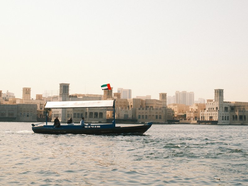 A traditional boat with a flag sailing on a calm waterway, surrounded by historic buildings.