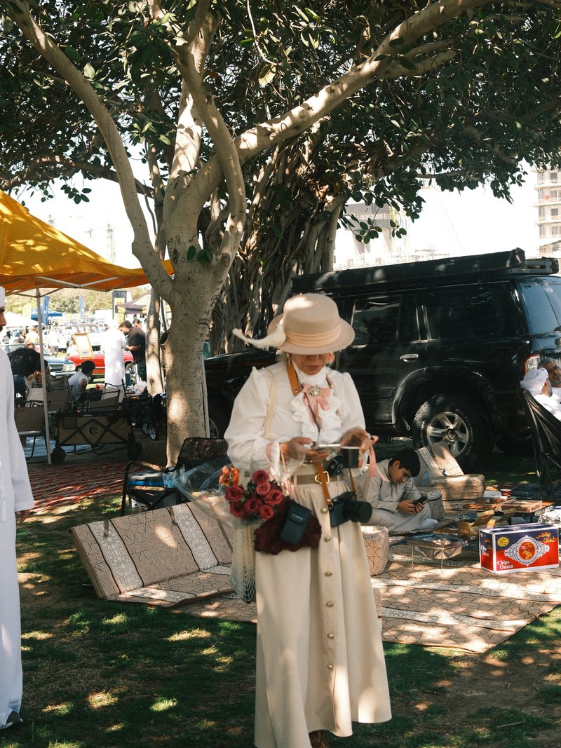 A woman in a white outfit and hat stands under a tree, holding a camera in a lively outdoor setting.