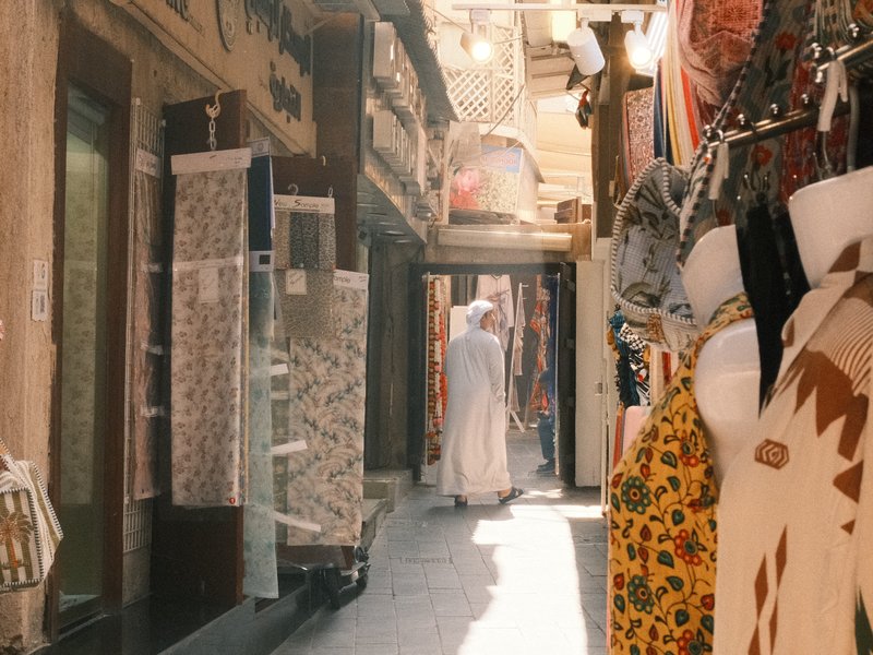 A narrow alleyway in a bustling market with colorful fabrics and a person in traditional attire walking away.