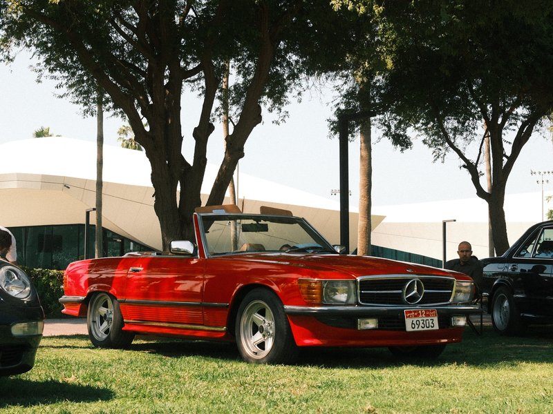 A red convertible Mercedes-Benz parked on green grass under trees with a modern building in the background.