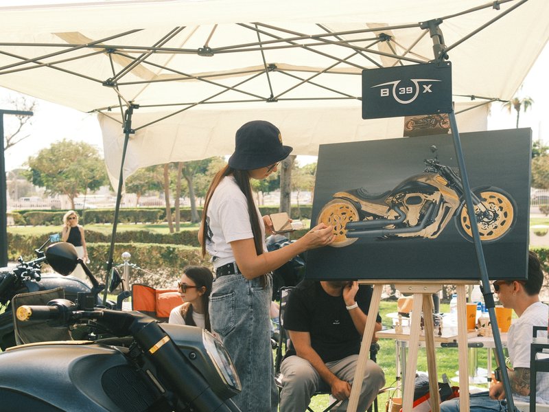 A woman painting a motorcycle on a canvas at an outdoor event with people and tents in the background.