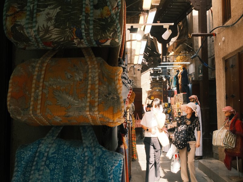 Colorful bags hanging in a narrow market alley with shoppers in the background.