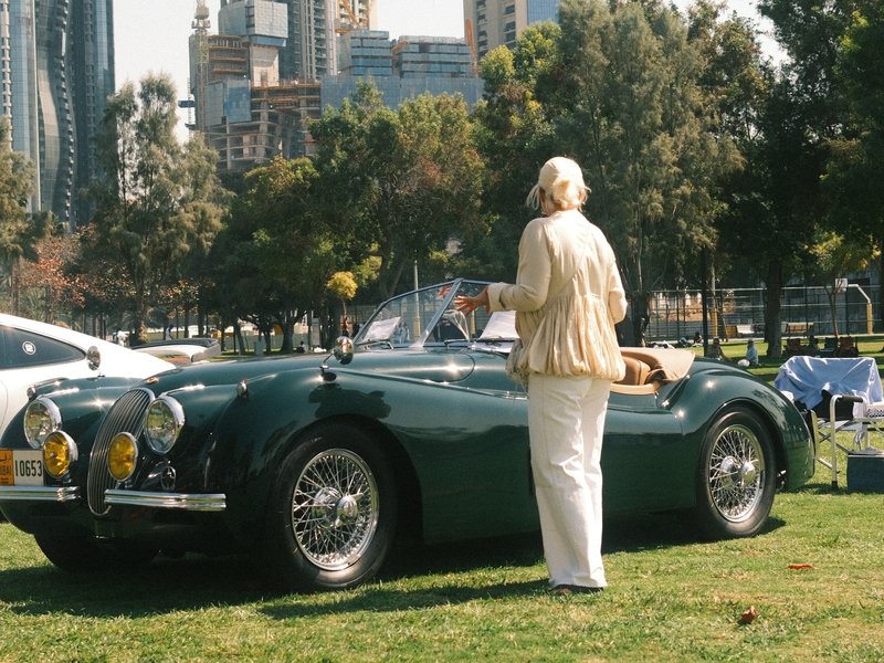 A vintage green car parked on grass with a person in light clothing standing beside it, city skyline in background.
