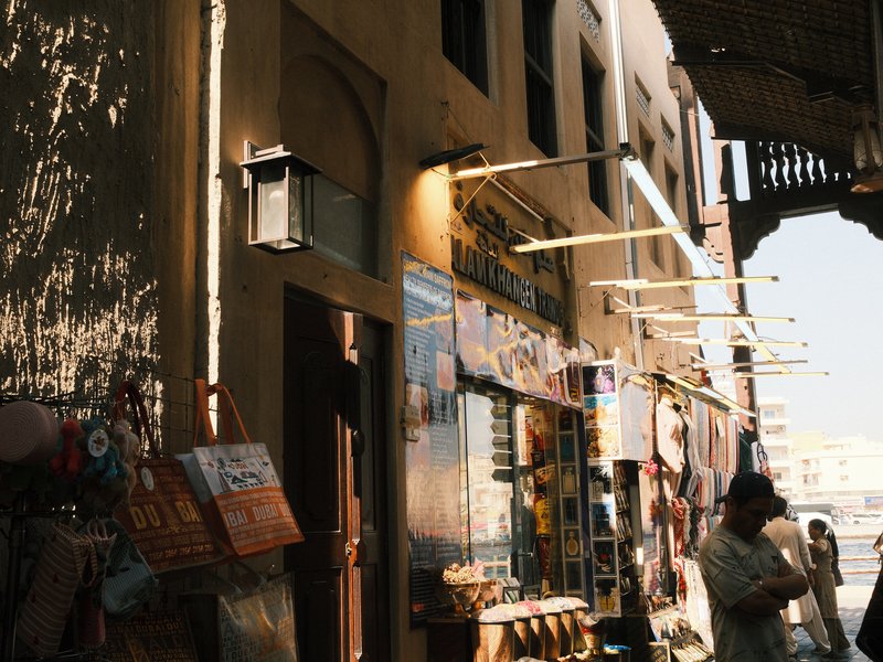 Narrow street with shops displaying colorful signs and merchandise, sunlight filtering through.