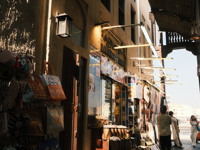 Narrow street lined with shops, sunlight illuminating the walls and people walking by.