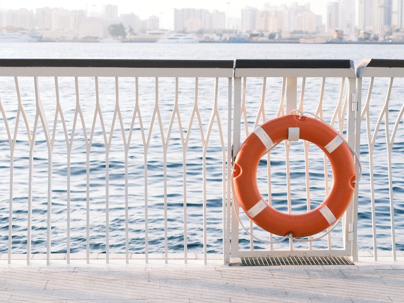 A bright orange life buoy hanging on a white railing with calm water and city skyline in the background.