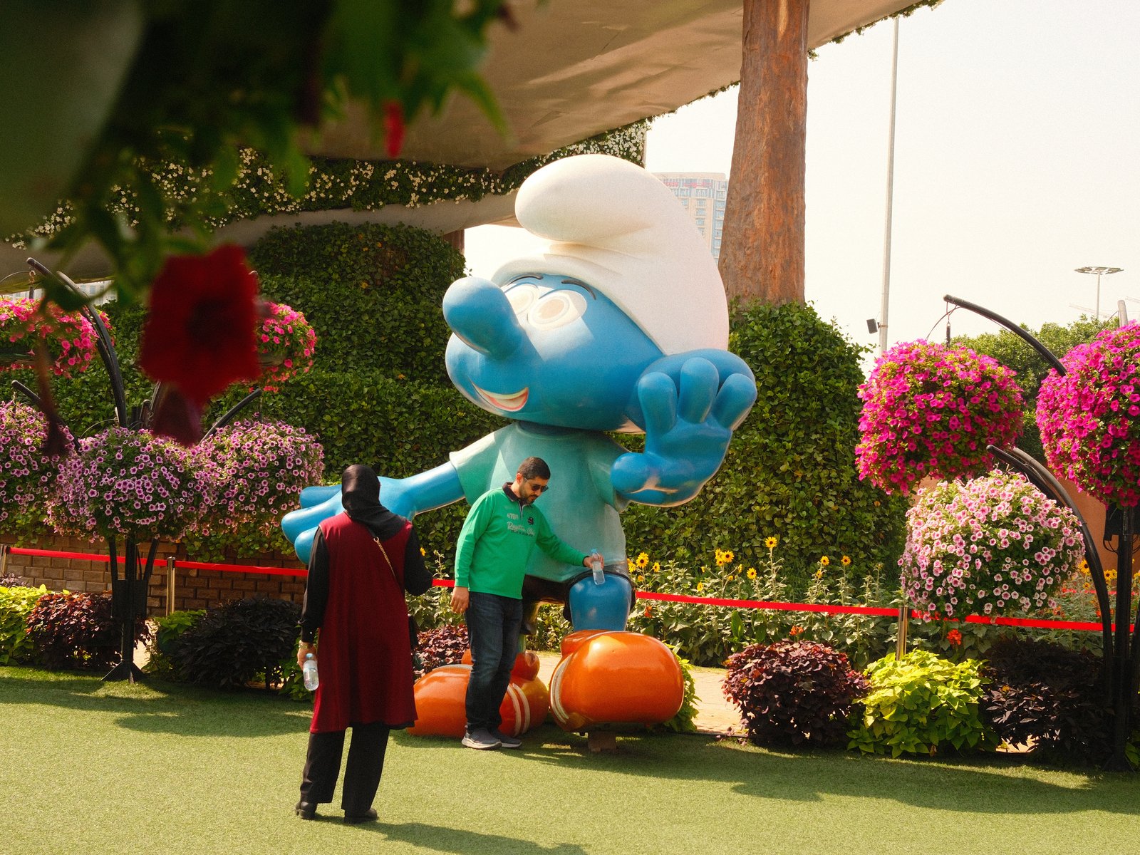 A large blue Smurf statue surrounded by colorful flowers and a person interacting with it.