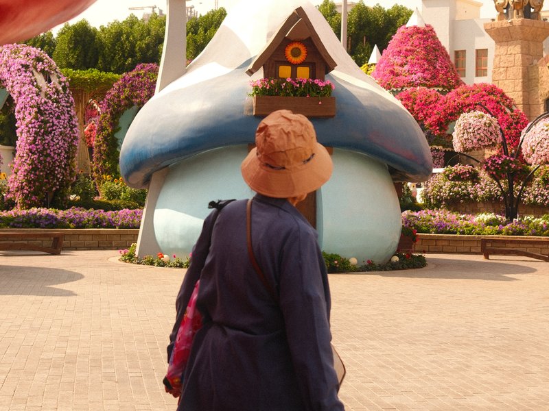 A person in a hat stands before a large mushroom-shaped structure surrounded by colorful flowers.