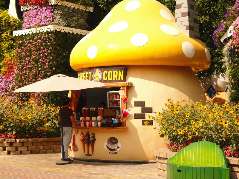 A whimsical mushroom-shaped food stand selling popcorn, surrounded by colorful flowers.
