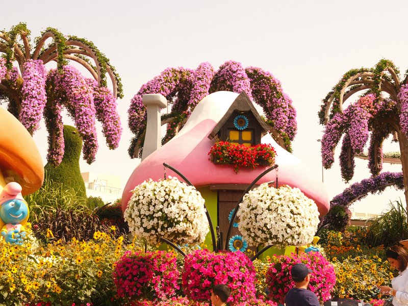 Colorful flower-covered house surrounded by vibrant floral displays and palm-like trees.