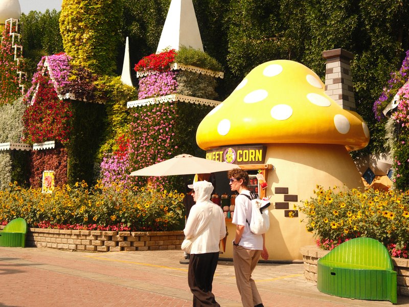 Two people walking past colorful mushroom-shaped structure in a flower-filled park.