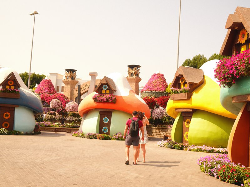 Two people walking past colorful mushroom-shaped houses adorned with flowers in a vibrant outdoor setting.