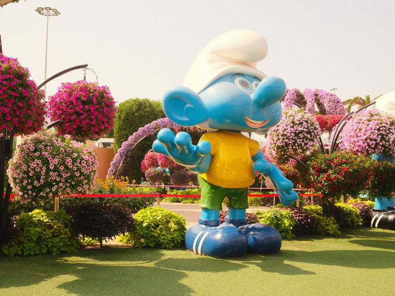 A large blue cartoon character statue in a flower garden, surrounded by colorful floral arrangements.