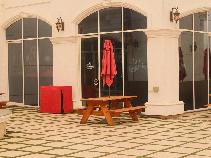 A patio area with wooden picnic tables, a red umbrella, and a red box against a backdrop of large glass windows.