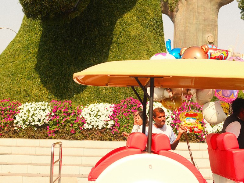 A person waving from a colorful cart in front of a large topiary and flower beds.