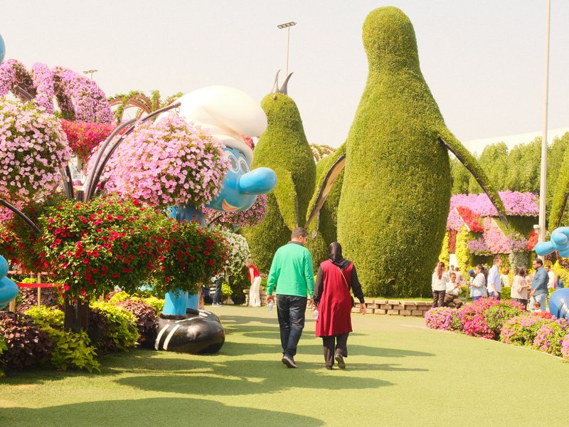 Two people walking through a vibrant flower garden with large topiary figures and colorful floral displays.