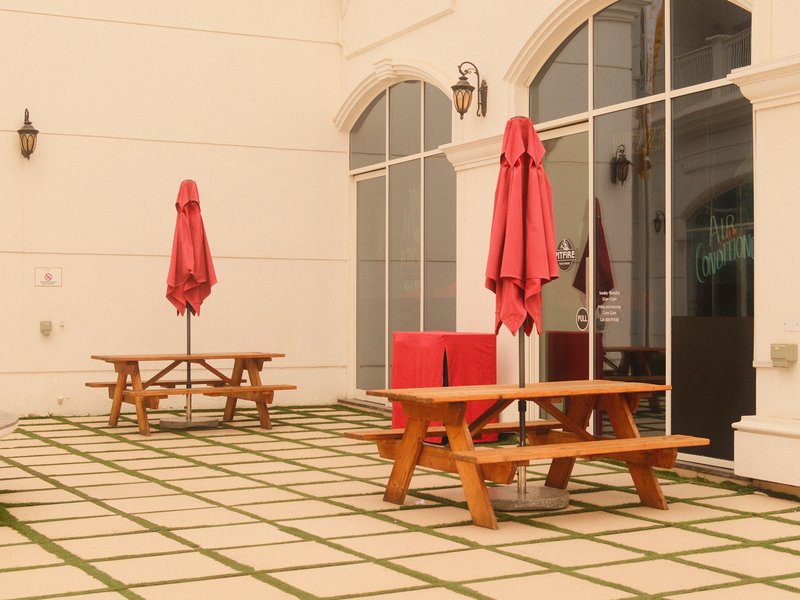 Two wooden picnic tables with red umbrellas on a tiled patio outside a building.