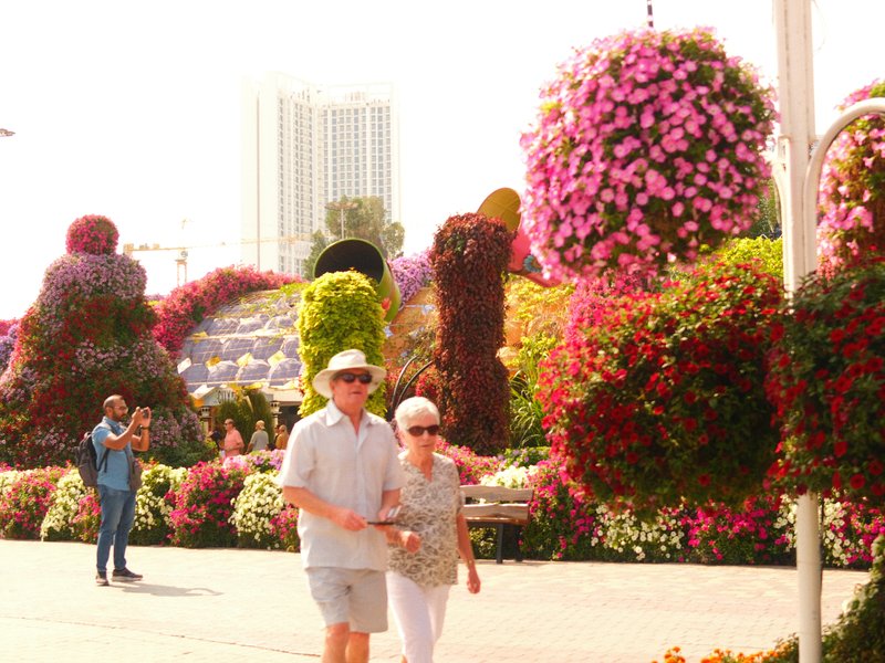 A couple walking hand-in-hand through a vibrant flower garden filled with hanging floral arrangements.