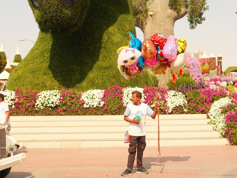 A young boy holding a stick with colorful balloons in front of a large topiary sculpture and flowers.