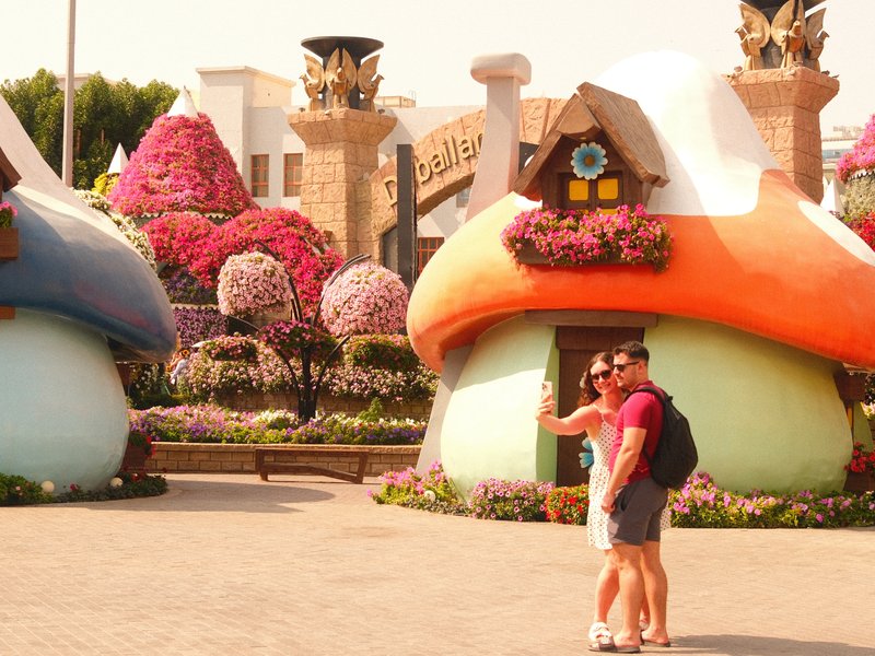 A couple poses for a selfie in front of colorful mushroom-shaped houses surrounded by vibrant flowers.