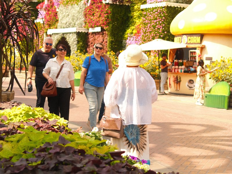 A group of people walking in a vibrant outdoor setting with colorful flowers and a food stall.