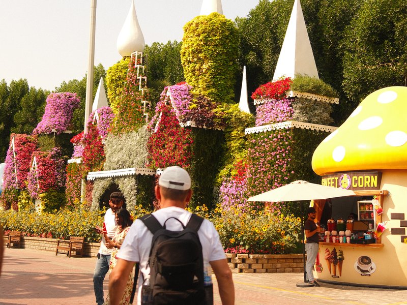 Colorful floral castle structure with people walking by and a food stall in a park.