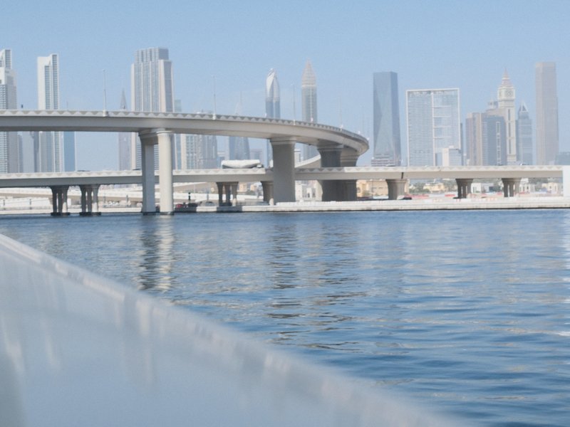 A view of a modern city skyline with a bridge over a calm water body under a clear blue sky.