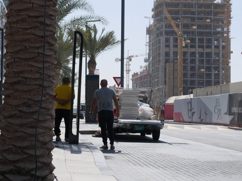 Two workers unloading materials from a truck on a sunny urban street with construction in the background.