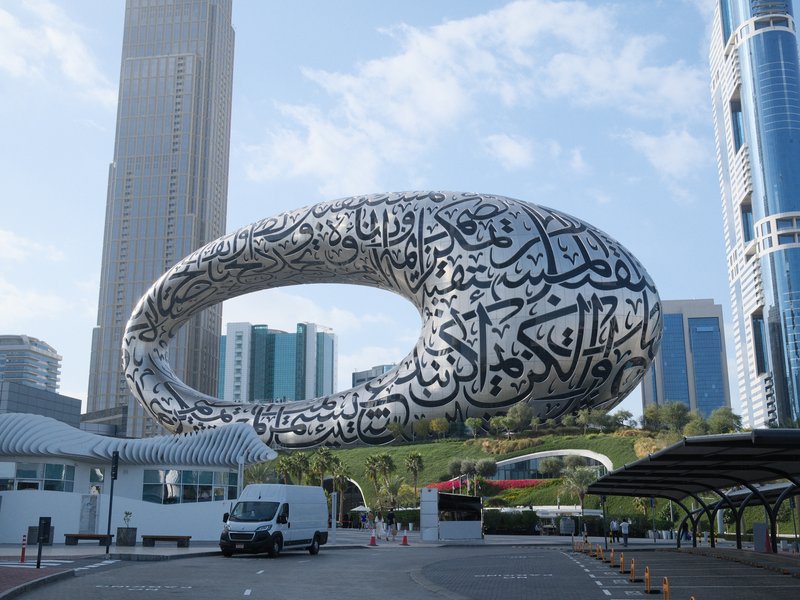 A modern architectural structure with Arabic calligraphy, surrounded by urban buildings and a clear sky.