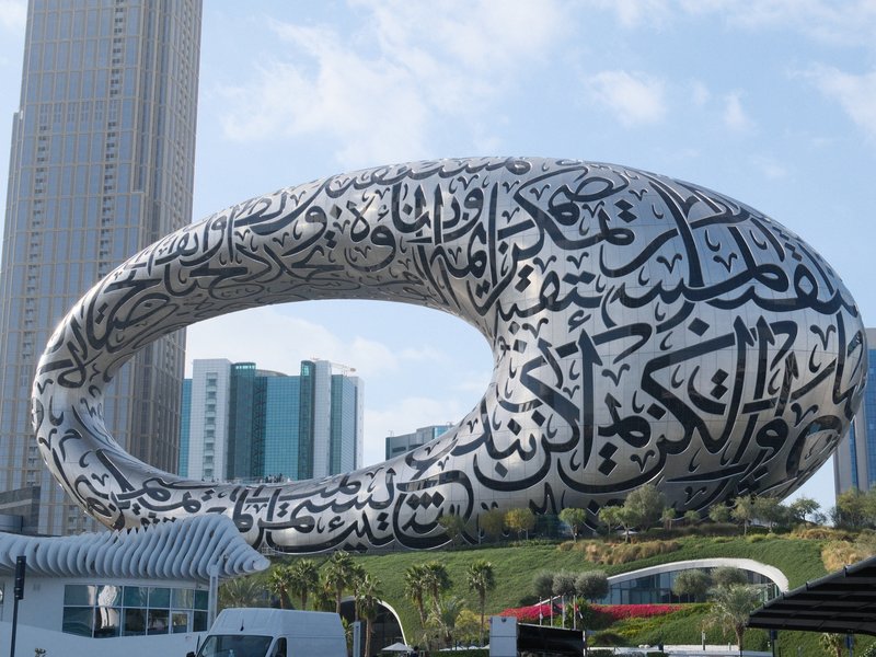 A large, modern sculpture featuring Arabic calligraphy, set against a city skyline.