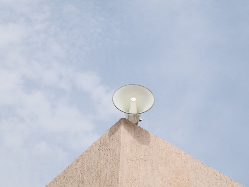 A white loudspeaker mounted on a beige wall against a blue sky with scattered clouds.