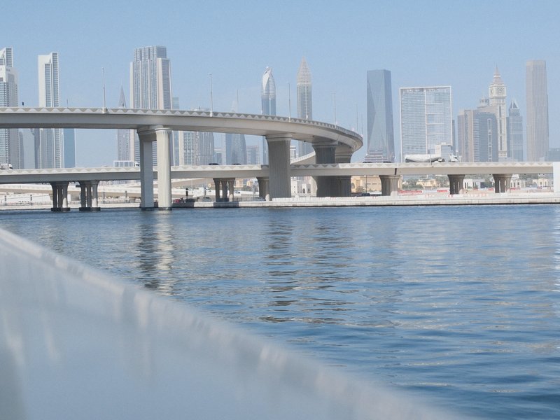 A modern city skyline with a curved bridge over calm water, reflecting skyscrapers under a clear blue sky.