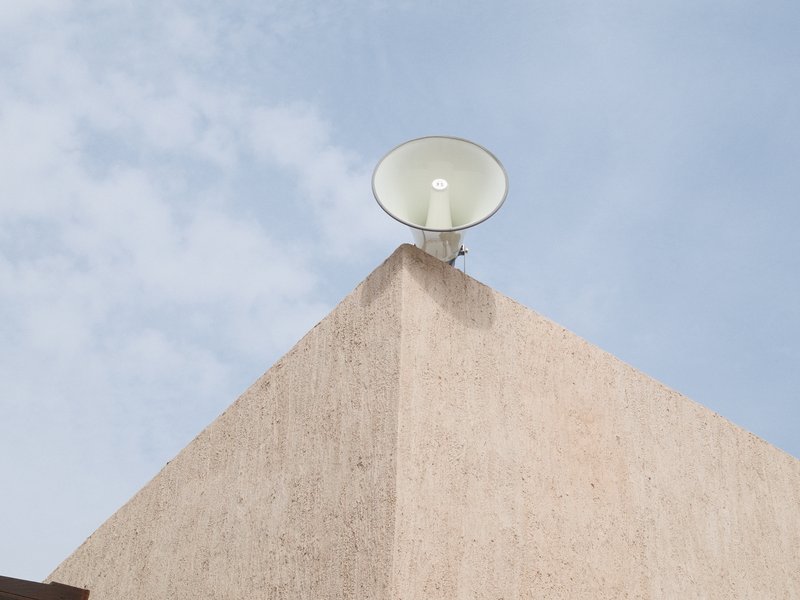 A white loudspeaker mounted on a beige wall against a blue sky with scattered clouds.