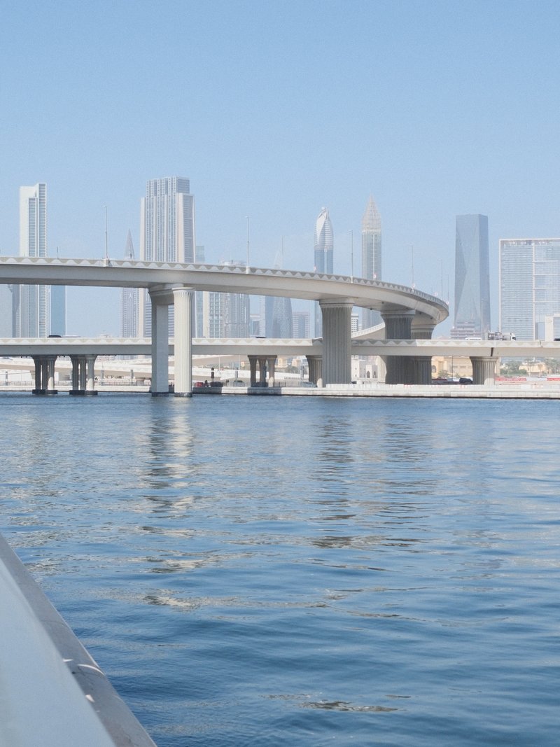 A modern city skyline with skyscrapers reflected in calm water, featuring a curved bridge.
