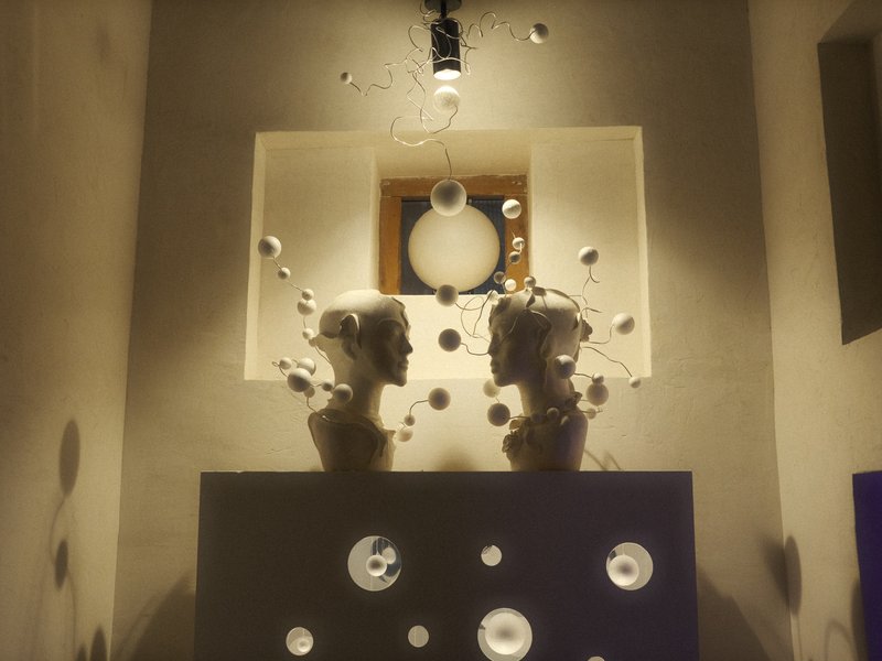 Two artistic busts on a dark pedestal surrounded by floating circular objects and a light fixture above.
