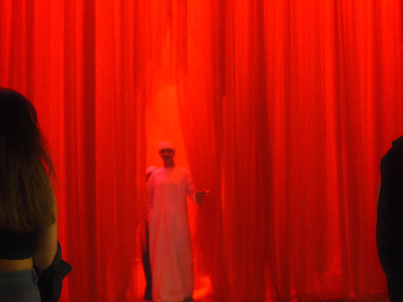 A figure in a white dress stands in front of a bright red curtain, creating a dramatic scene.