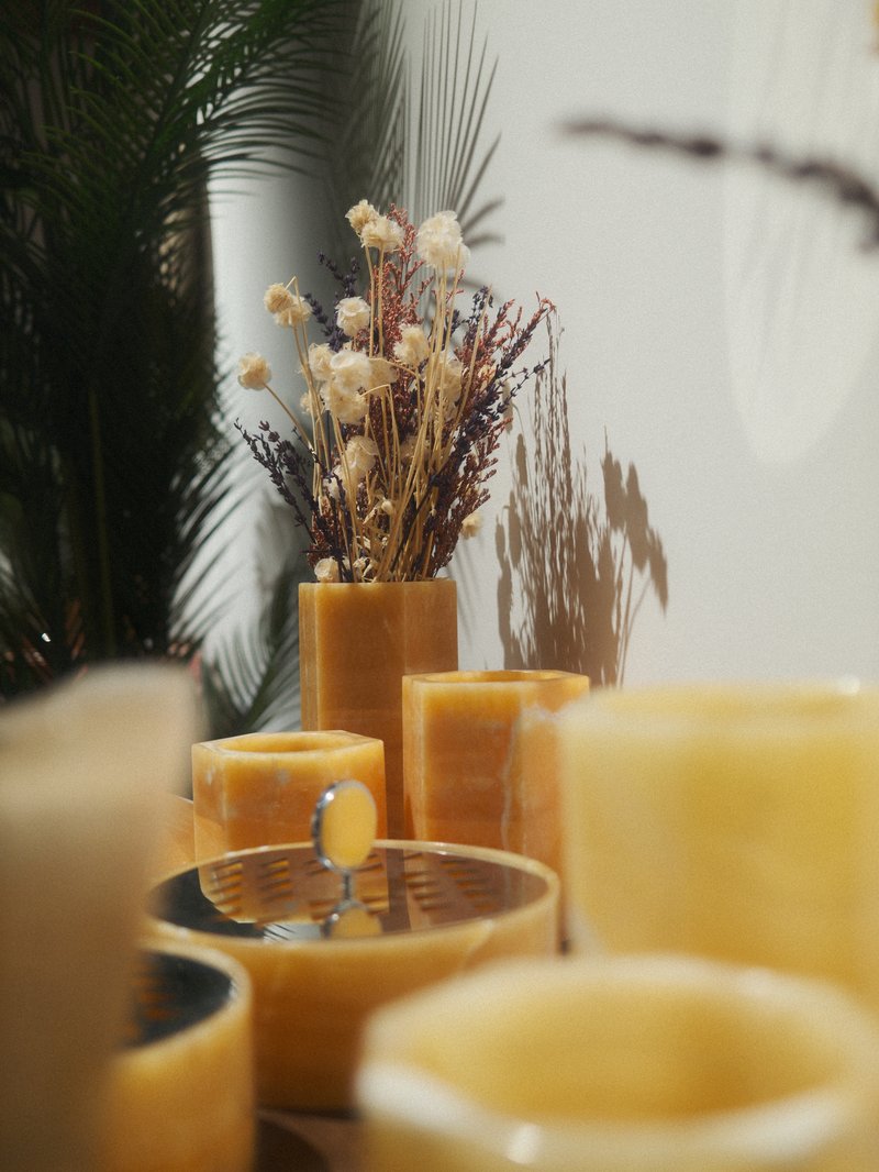 A collection of yellow candles and a decorative vase with dried flowers against a light background.