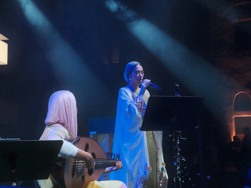 Two female musicians performing on stage, one singing and the other playing an instrument, with dramatic lighting.