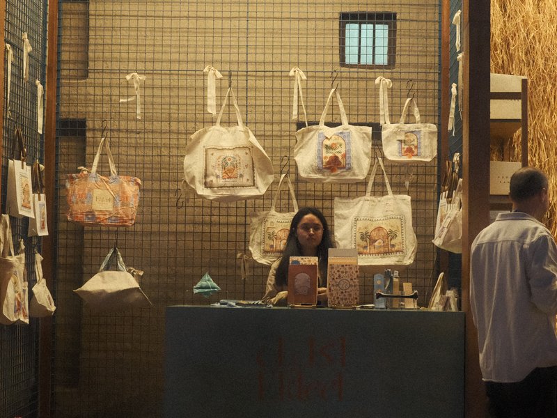 A woman stands behind a counter in a shop displaying tote bags on a grid wall.