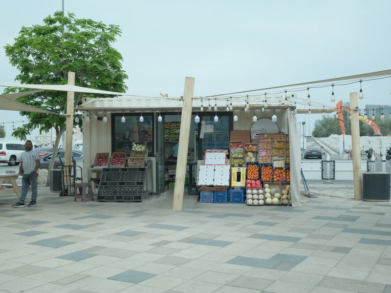 A small market stall with colorful produce and crates, set in a plaza with a cloudy sky.