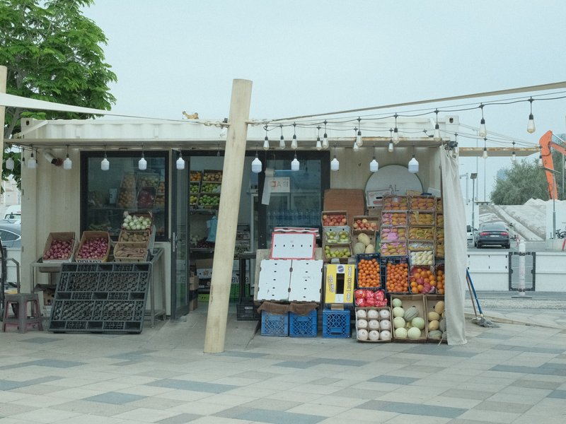 A fruit and vegetable stall with colorful produce displayed outside a small shop under a cloudy sky.
