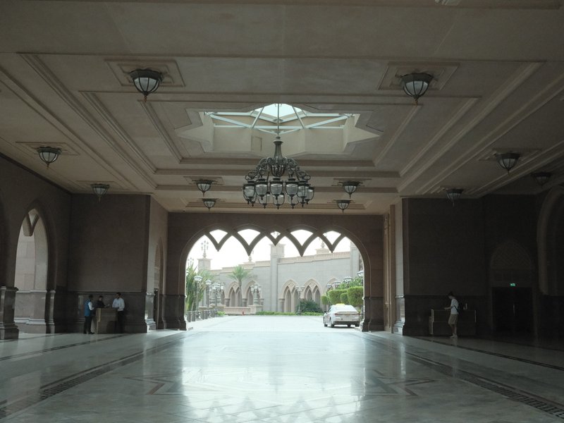 Interior view of a grand hall with arches and chandeliers, leading to an outdoor courtyard.