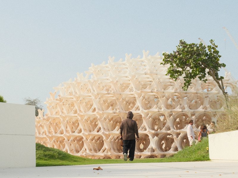 A person walking towards a large, intricate architectural structure made of interlocking shapes, surrounded by greenery.