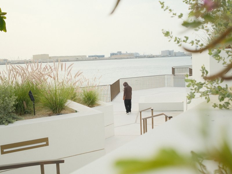 A solitary figure walks along a white pathway by the water, surrounded by greenery and soft light.