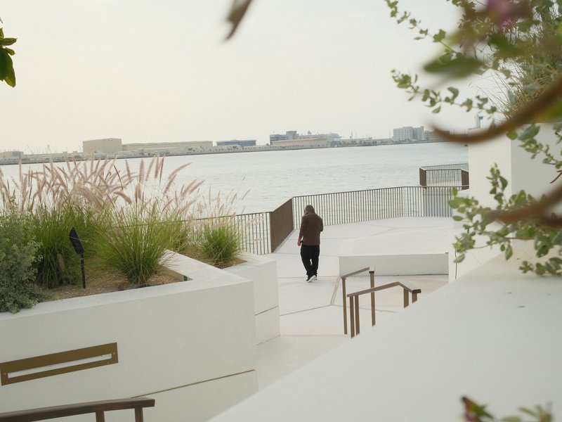 A person walking along a white pathway by the water, surrounded by greenery and distant buildings.