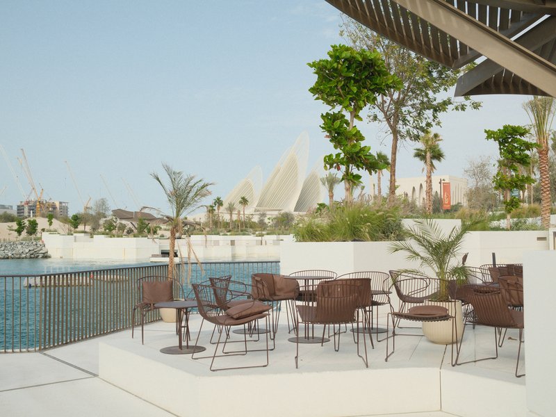 Outdoor seating area by a water body with modern architecture in the background and greenery.